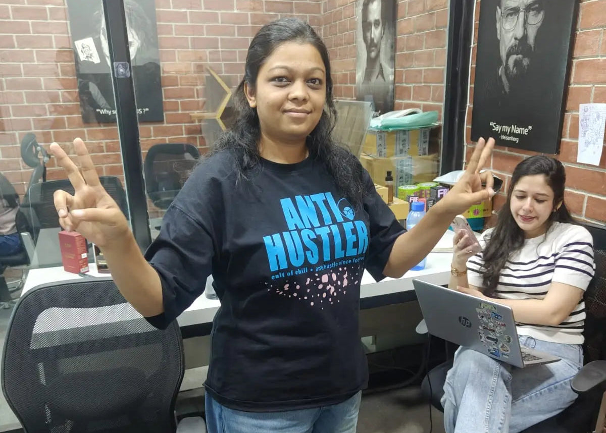 A woman standing in an office wearing a black graphic t-shirt with 'ANTI-HUSTLER' printed in blue and pink, paired with blue jeans. 