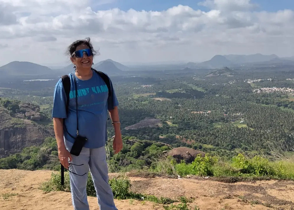 A woman wearing a blue Bahut Kaam Bahut Calm t-shirt, light gray pants, and sunglasses stands on a rocky outcrop overlooking a vast green valley and distant mountains under a cloudy sky.