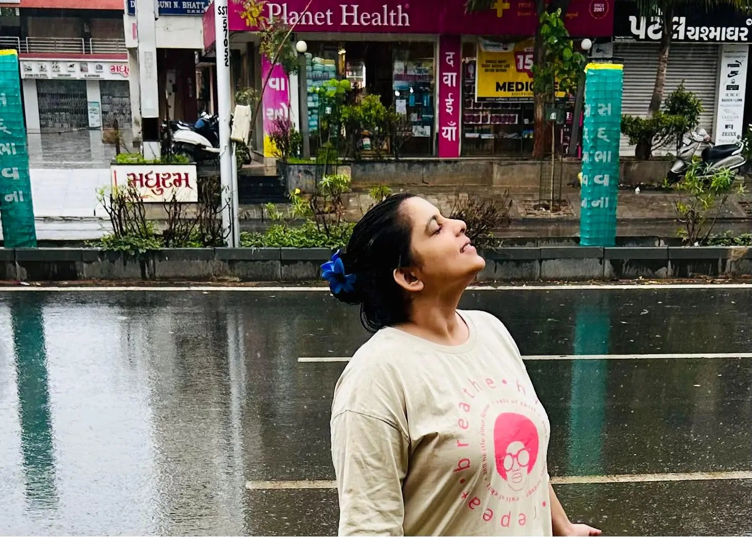 A woman wearing a beige t-shirt with a pink graphic Breathe Hydrate Repeat print stands on a wet city street, looking upward.