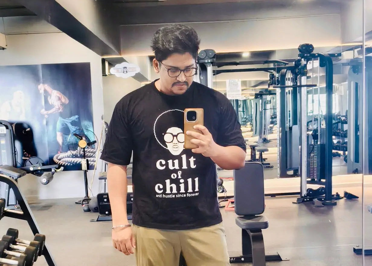 A man taking a mirror selfie in a gym, wearing a black graphic t-shirt with white text that reads 'Cult of Chill' and khaki pants.