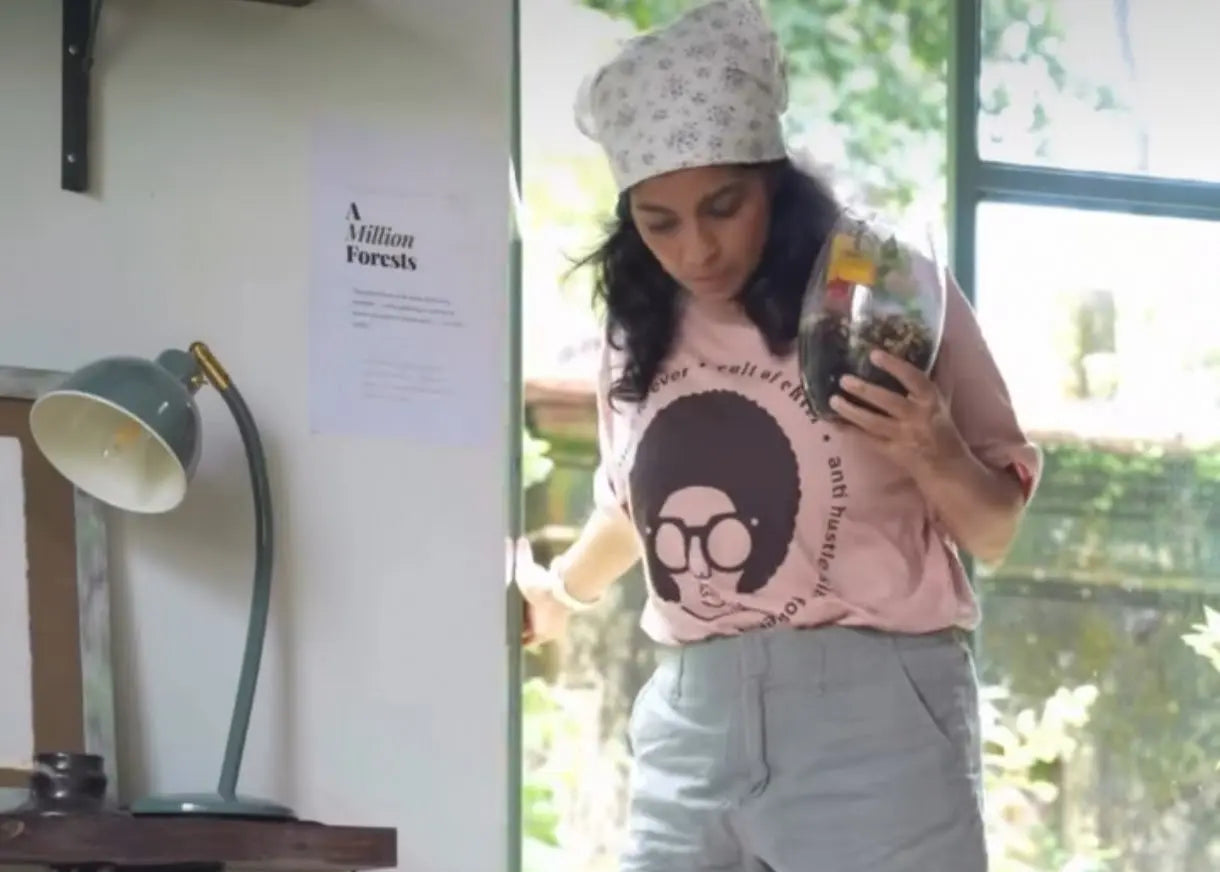 A woman stands in a bright room near a window, holding a small glass jar. She is wearing a light pink graphic T-shirt with a black-and-white afro illustration and the text Cult of Chill, light gray shorts, and a white patterned beanie.