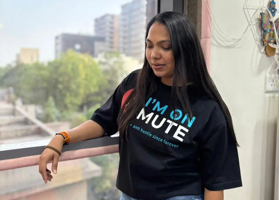 A woman wearing a black graphic t-shirt with the text 'I'M ON MUTE' in blue and white, paired with light blue jeans, standing by a window with a city view.