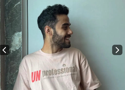 A man wearing a light pink t-shirt with 'UNprofessional' printed on the front, standing against a pale blue-gray wall.